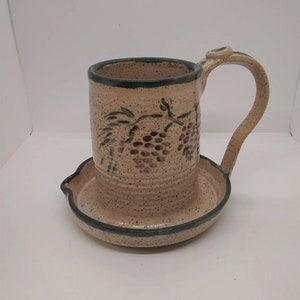 May include: A beige ceramic mug with a green rim and a green vine design with brown grapes. The mug has a matching saucer with a green rim.