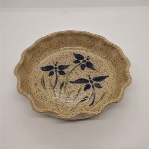 May include: A beige ceramic bowl with a speckled finish and a wavy rim. The bowl is decorated with three blue floral designs and green stems. This decorative bowl is ideal for serving or display.