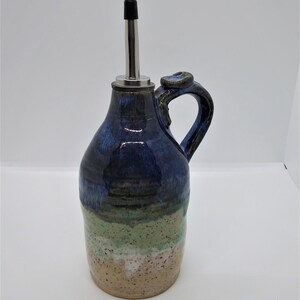 May include: A handmade ceramic olive oil bottle with a blue, green, and beige glaze. The bottle has a spout with a metal top and a handle.