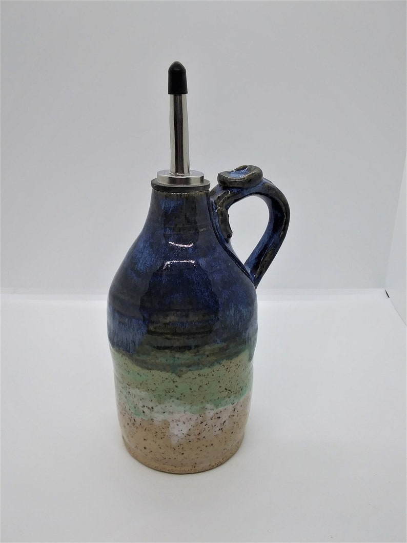 May include: A handmade ceramic olive oil bottle with a blue, green, and beige glaze. The bottle has a spout with a metal top and a handle.