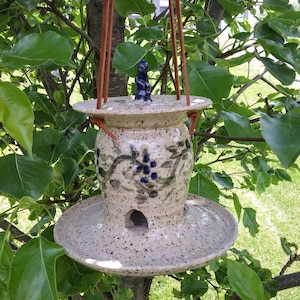 May include: A speckled ceramic bird feeder with a round base and a small opening. The feeder has a decorative blue floral design and is suspended by brown leather straps. A small blue figurine sits on top.