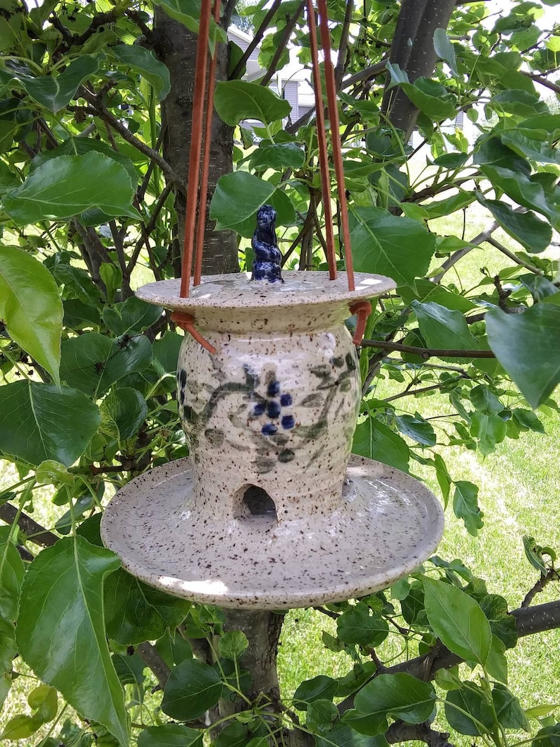 May include: A speckled ceramic bird feeder with a round base and a small opening. The feeder has a decorative blue floral design and is suspended by brown leather straps. A small blue figurine sits on top.