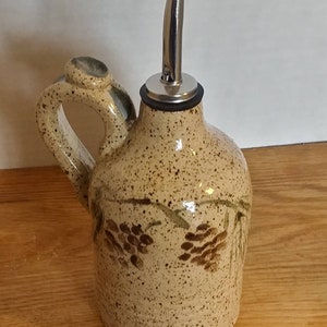 May include: A ceramic olive oil bottle with a brown speckled glaze and a pine cone design. The bottle has a metal pour spout and a handle.