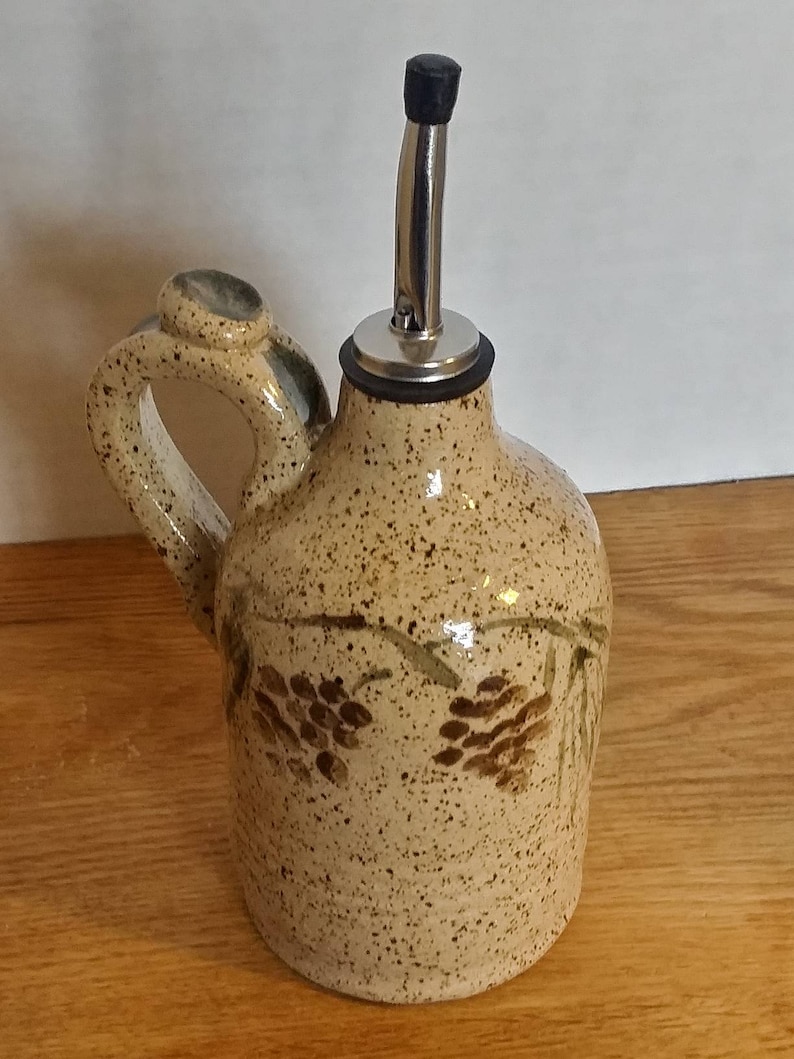 May include: A ceramic olive oil bottle with a brown speckled glaze and a pine cone design. The bottle has a metal pour spout and a handle.