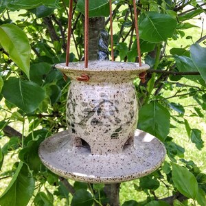 May include: A speckled ceramic bird feeder with a lid and a circular base, suspended by brown leather straps. The feeder has a textured surface with green and brown accents. The feeder is hanging from a tree branch.