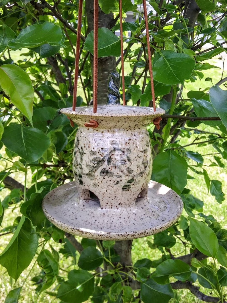 May include: A speckled ceramic bird feeder with a lid and a circular base, suspended by brown leather straps. The feeder has a textured surface with green and brown accents. The feeder is hanging from a tree branch.