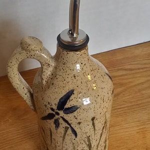 May include: A ceramic olive oil bottle with a speckled brown glaze and a dragonfly design. The bottle has a metal pour spout and a handle.