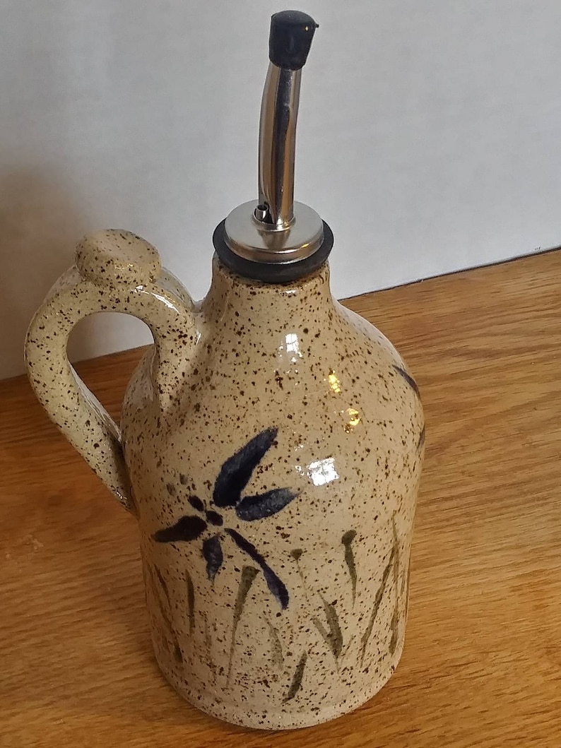 May include: A ceramic olive oil bottle with a speckled brown glaze and a dragonfly design. The bottle has a metal pour spout and a handle.