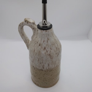 May include: A white ceramic oil and vinegar bottle with a brown speckled glaze. The bottle has a handle and a stainless steel pour spout with a black rubber stopper.