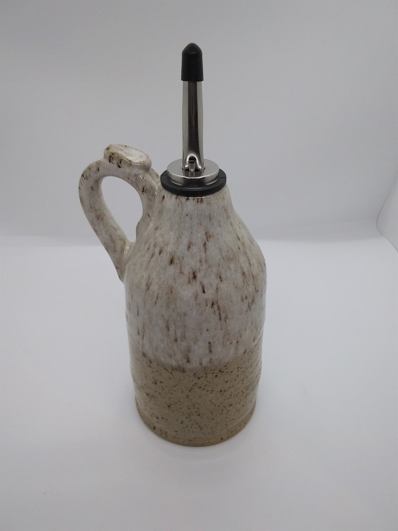 May include: A white ceramic oil and vinegar bottle with a brown speckled glaze. The bottle has a handle and a stainless steel pour spout with a black rubber stopper.