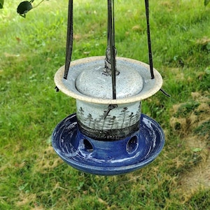 May include: A handcrafted ceramic bird feeder with a speckled gray lid and a blue base. The feeder has a cylindrical body with a forest silhouette design and is suspended by black metal wires. The feeder is hanging outdoors against a backdrop of green grass.