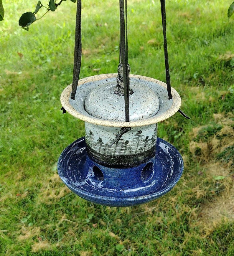 May include: A handcrafted ceramic bird feeder with a speckled gray lid and a blue base. The feeder has a cylindrical body with a forest silhouette design and is suspended by black metal wires. The feeder is hanging outdoors against a backdrop of green grass.