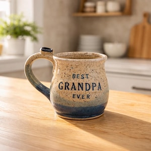 May include: A ceramic mug with a speckled beige finish, featuring the text "BEST GRANDPA EVER" in navy blue. The mug has a blue gradient at the base and on the handle, and sits on a wooden surface.