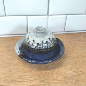 Könnte beinhalten: Eine handgefertigte Keramik-Butterdose mit Deckel. Die Schale ist tiefblau und der Deckel hellgrau mit einem schwarz-weißen Waldmuster. Die Schale steht auf einer Holzoberfläche.