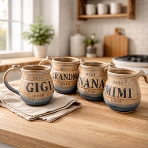 Op de afbeelding: Vier keramische mokken met handvatten, elk met een andere naam: "GIGI", "GRANDMA", "NANA" en "MIMI". De mokken hebben een beige en blauw kleurenschema met de woorden "BEST EVER" boven en onder de namen.