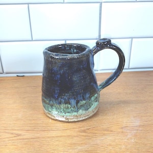 Puede incluir: Taza de cerámica hecha a mano con un esmalte azul oscuro que se desvanece a turquesa y blanco cerca de la base. La taza tiene un asa redondeada y una apariencia texturizada única. Una pieza funcional y decorativa.