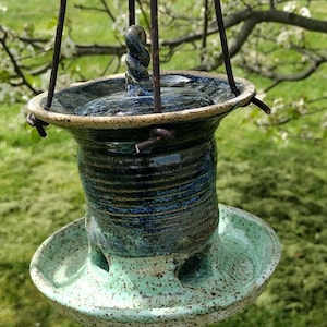 May include: Handmade ceramic bird feeder with a speckled turquoise base and a dark blue and brown body. The feeder has a circular design with a twisted handle and is suspended by metal rods.