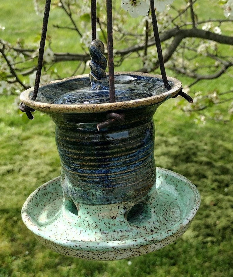 May include: Handmade ceramic bird feeder with a speckled turquoise base and a dark blue and brown body. The feeder has a circular design with a twisted handle and is suspended by metal rods.