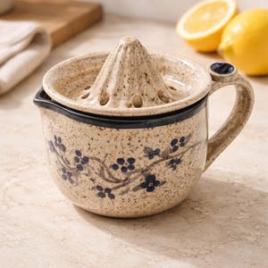 Puede incluir: Exprimidor de cítricos de cerámica con acabado beige moteado y diseño floral azul oscuro. El exprimidor tiene un escariador incorporado y un asa. Dos mitades de limón en el fondo.