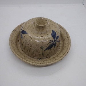 May include: A ceramic butter dish with a lid. The dish is speckled brown and beige with blue floral designs. The lid has a small opening in the center.