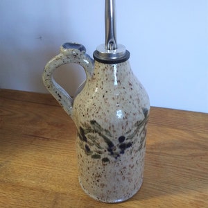 May include: A ceramic olive oil bottle with a speckled glaze and a floral design. The bottle has a spout with a metal cap and a handle.