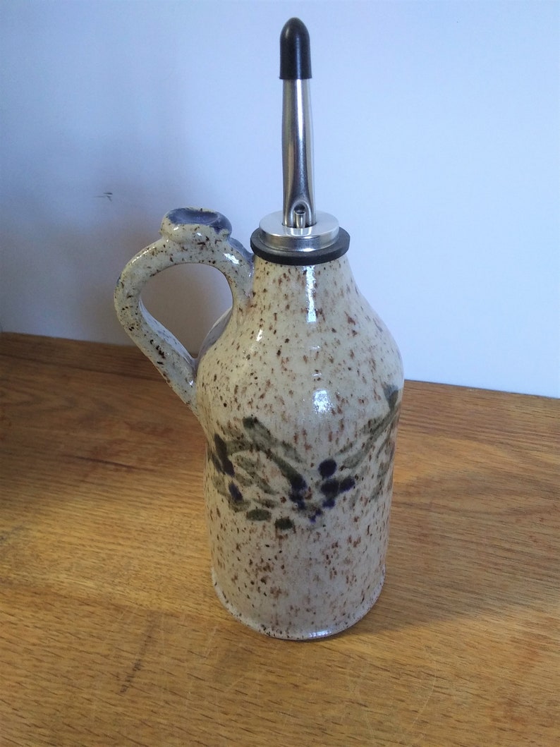 May include: A ceramic olive oil bottle with a speckled glaze and a floral design. The bottle has a spout with a metal cap and a handle.