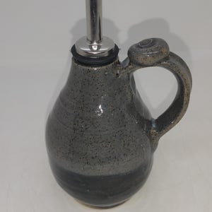 May include: A gray ceramic oil and vinegar bottle with a black pour spout and a loop handle. The bottle has a speckled glaze and a dark gray band around the bottom.