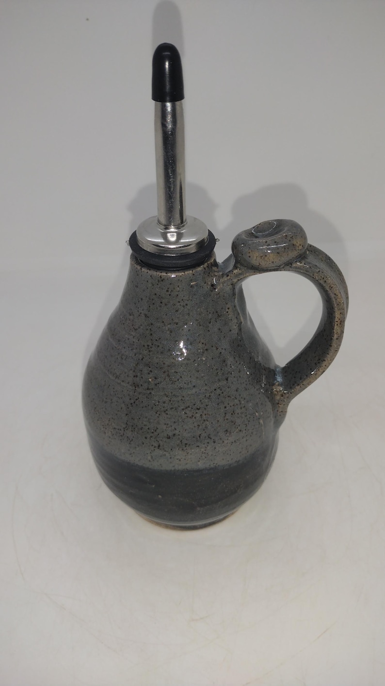 May include: A gray ceramic oil and vinegar bottle with a black pour spout and a loop handle. The bottle has a speckled glaze and a dark gray band around the bottom.