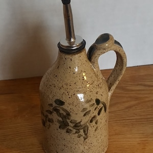 May include: A ceramic olive oil bottle with a brown speckled glaze and green leaf design. The bottle has a metal pour spout and a handle.
