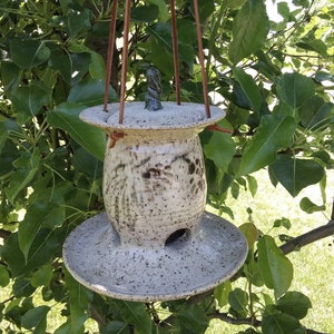 May include: A speckled ceramic bird feeder with a round base and top, suspended by brown leather straps. The feeder has a cylindrical body with a small opening and a decorative top. The background features green leaves and branches.