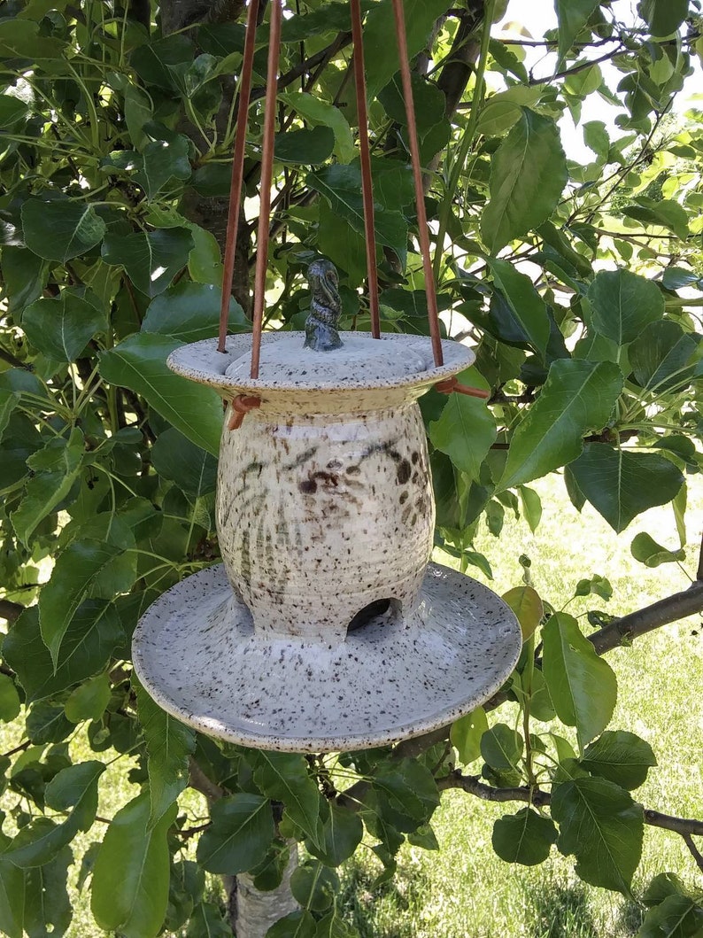 May include: A speckled ceramic bird feeder with a round base and top, suspended by brown leather straps. The feeder has a cylindrical body with a small opening and a decorative top. The background features green leaves and branches.