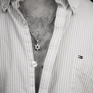 May include: A close-up of a man's chest with a silver chain necklace and a star of David pendant. The man is wearing a white striped button-down shirt.