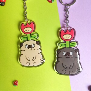 Puede incluir: Dos llaveros con un pug de dibujos animados con una flor que le crece de la cabeza. Un pug es beige y el otro es marrón. Ambas flores son rojas con una cara amarilla.