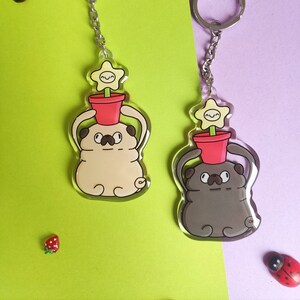Puede incluir: Dos llaveros con un pug de dibujos animados con una maceta en la cabeza. Un pug es marrón y el otro es beige. Ambos llaveros tienen cadenas y anillas plateadas.