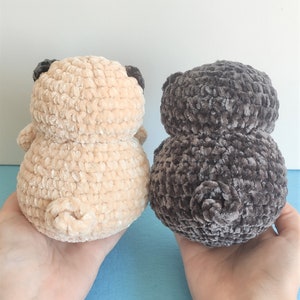 May include: Two crocheted pug dog plushies, one in beige and one in dark gray, are shown from behind. The plushies are made with a soft, fuzzy yarn.
