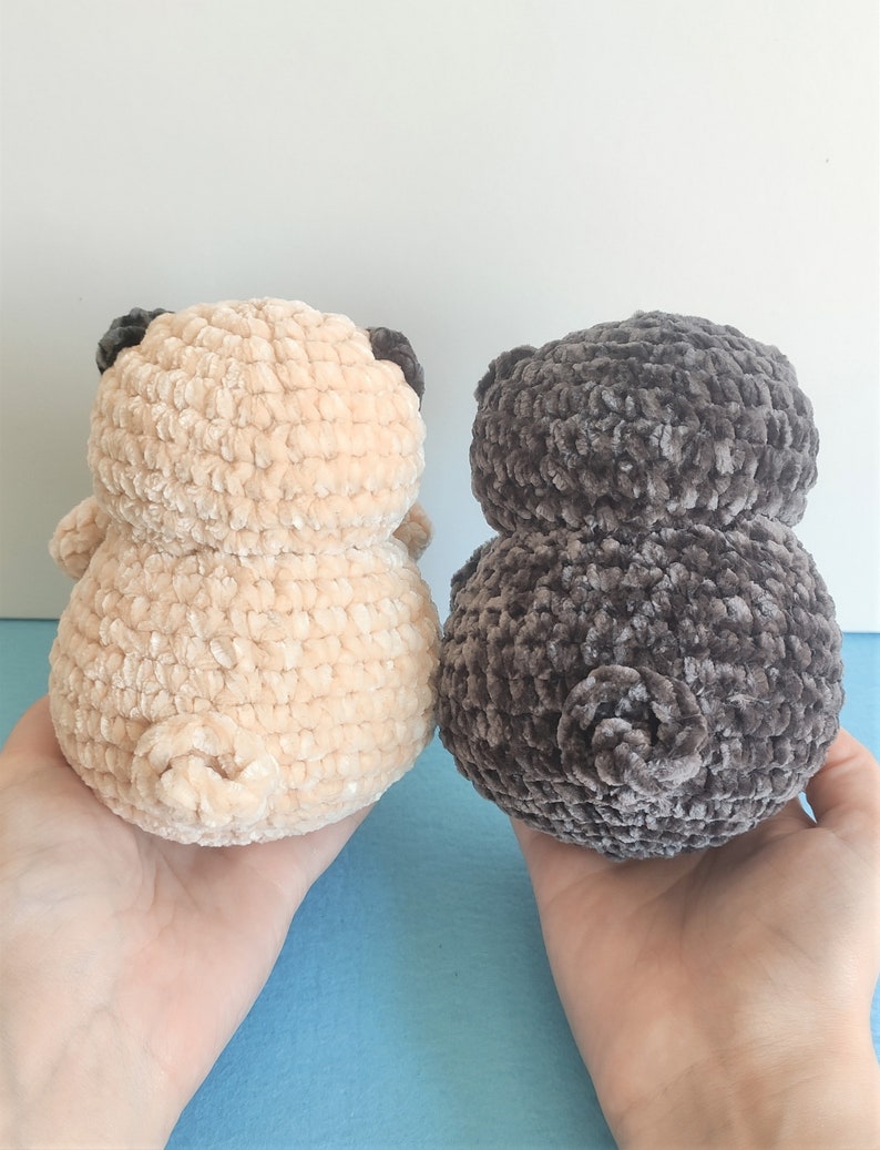 May include: Two crocheted pug dog plushies, one in beige and one in dark gray, are shown from behind. The plushies are made with a soft, fuzzy yarn.