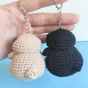 May include: Two crocheted keychains, one beige and one black, shaped like a pug dog. The keychains are hanging from silver chains.