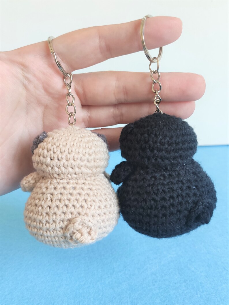 May include: Two crocheted keychains, one beige and one black, shaped like a pug dog. The keychains are hanging from silver chains.