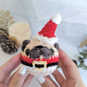Pu&ograve; includere: Un pug all'uncinetto che indossa un cappello da Babbo Natale rosso e bianco con una cintura da Babbo Natale nera e bianca. Il pug tiene un campanello giallo in bocca.