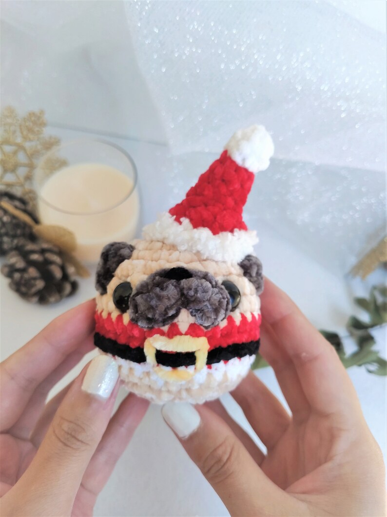 Pu&ograve; includere: Un pug all'uncinetto che indossa un cappello da Babbo Natale rosso e bianco con una cintura da Babbo Natale nera e bianca. Il pug tiene un campanello giallo in bocca.