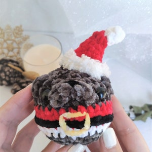 Pu&ograve; includere: Un giocattolo di peluche a forma di carlino lavorato a uncinetto che indossa un cappello da Babbo Natale rosso e bianco con una cintura nera e rossa. Il giocattolo ha occhi neri e una pelliccia grigia e bianca.