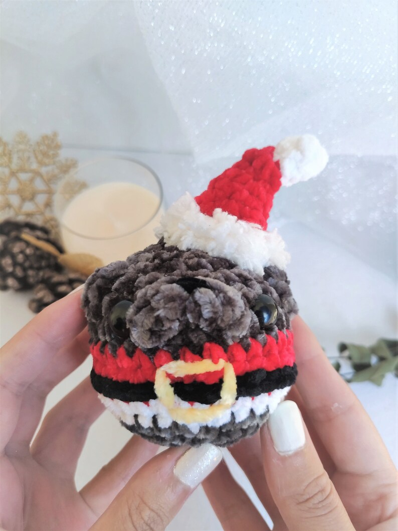 Pu&ograve; includere: Un giocattolo di peluche a forma di carlino lavorato a uncinetto che indossa un cappello da Babbo Natale rosso e bianco con una cintura nera e rossa. Il giocattolo ha occhi neri e una pelliccia grigia e bianca.