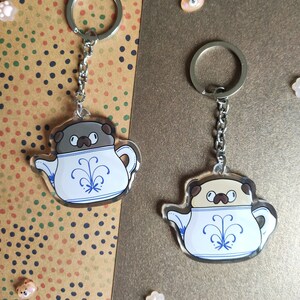 Pode incluir: Dois chaveiros de xícara de chá com um pug. Um chaveiro apresenta um pug marrom e o outro apresenta um pug marrom claro. Ambos os chaveiros têm um design azul e branco na xícara de chá.