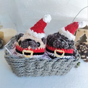 Pu&ograve; includere: Due ornamenti di pug in crochet che indossano cappelli di Babbo Natale e sciarpe rosse e nere in un cesto di vimini grigio.