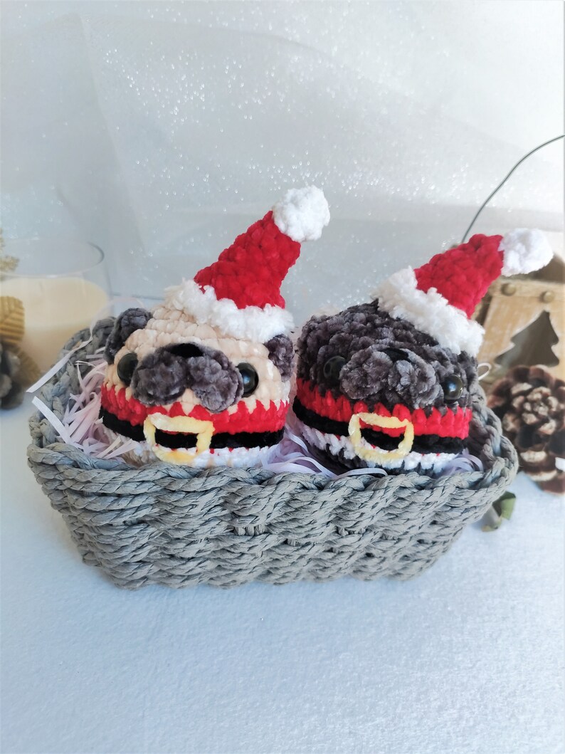 Pu&ograve; includere: Due ornamenti di pug in crochet che indossano cappelli di Babbo Natale e sciarpe rosse e nere in un cesto di vimini grigio.