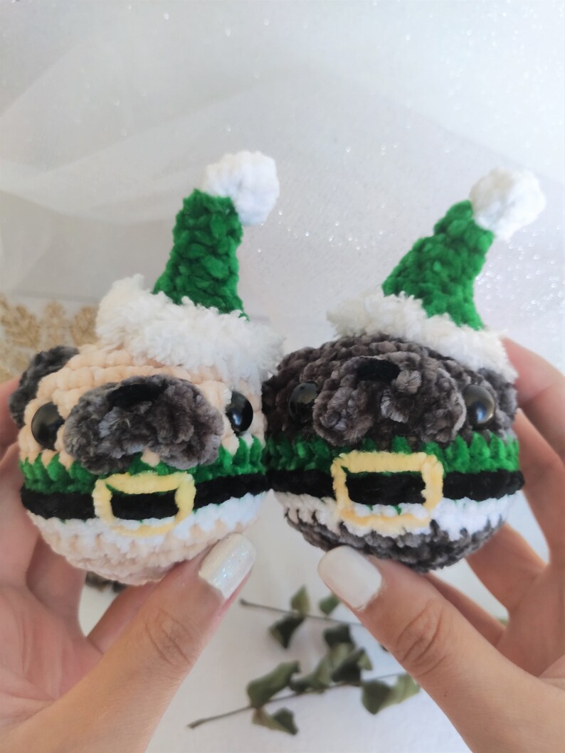 Pu&ograve; includere: Due pugs di peluche all'uncinetto che indossano cappelli di Babbo Natale verdi e bianchi con cinture nere e gialle.