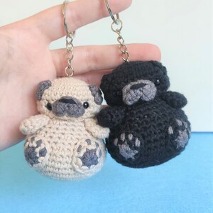 May include: Two crocheted pug dog keychains, one beige and one black, with gray paw prints. The keychains are attached to silver key rings.