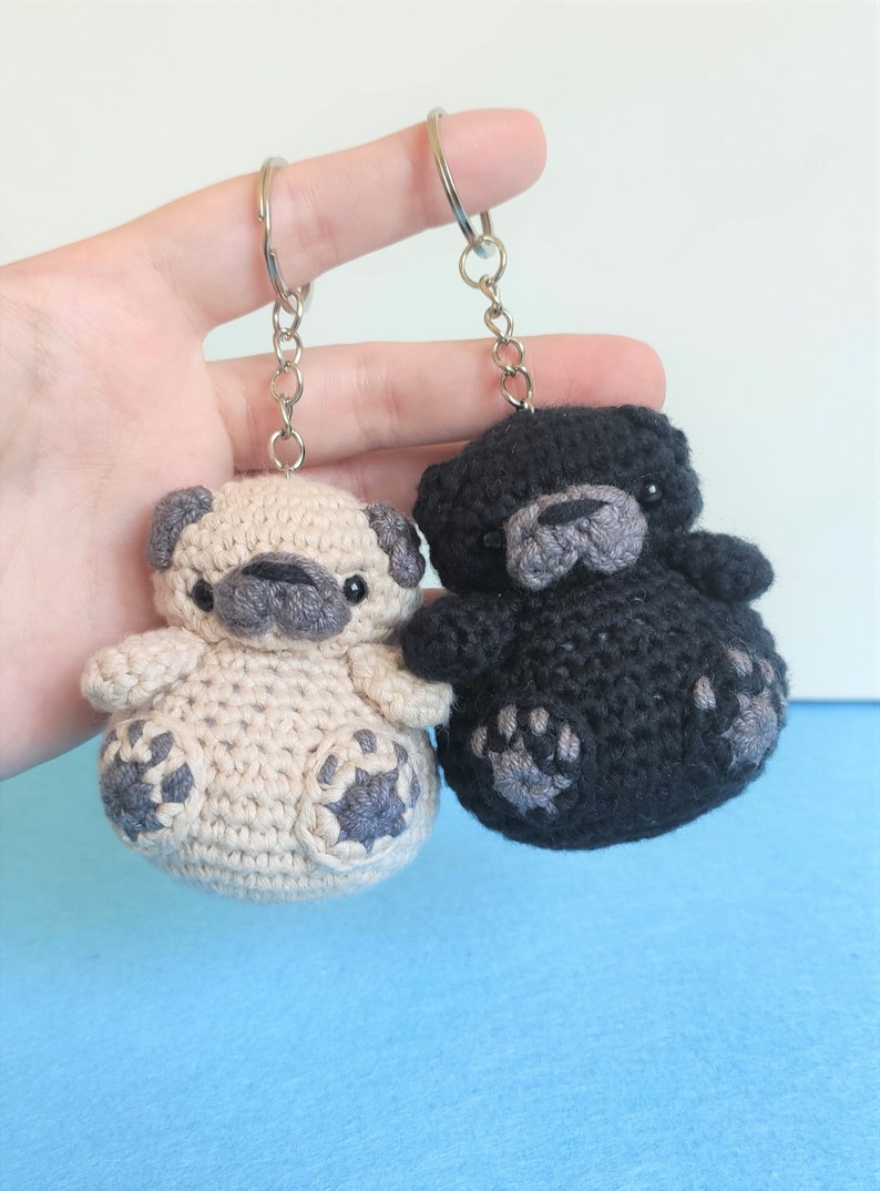 May include: Two crocheted pug dog keychains, one beige and one black, with gray paw prints. The keychains are attached to silver key rings.