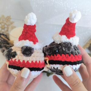 Pu&ograve; includere: Due giocattoli di peluche all'uncinetto a forma di carlino che indossano cappelli da Babbo Natale. I carlini indossano sciarpe a righe rosse e nere. Un carlino &egrave; beige e l'altro &egrave; grigio.