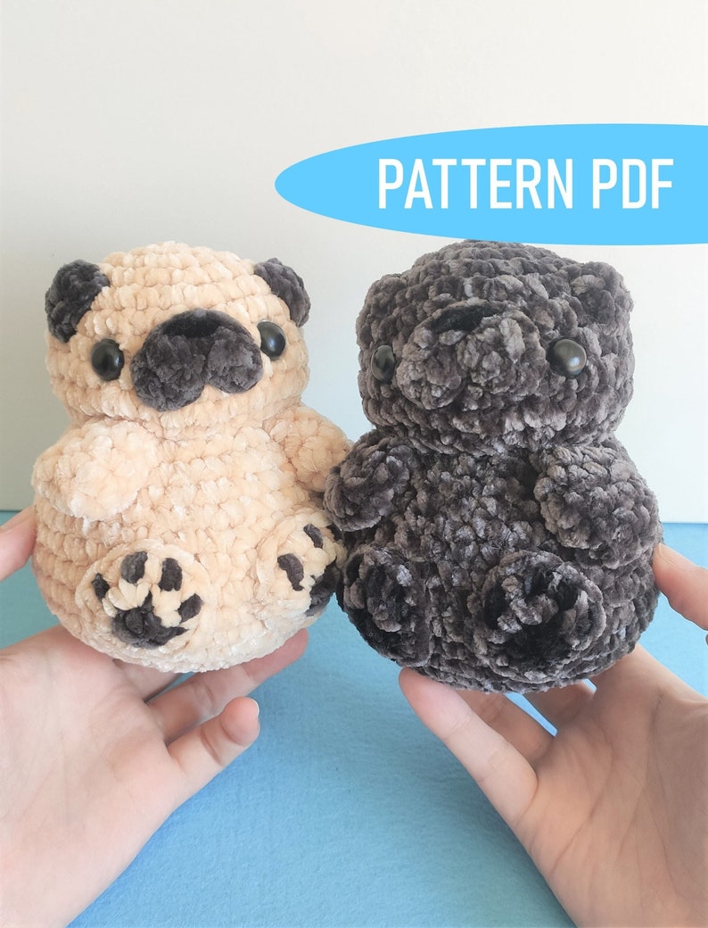 May include: Two crocheted plush pug dog toys. One is beige with brown accents and the other is dark gray.  The toys are being held in the hands of a person.  PATTERN PDF.